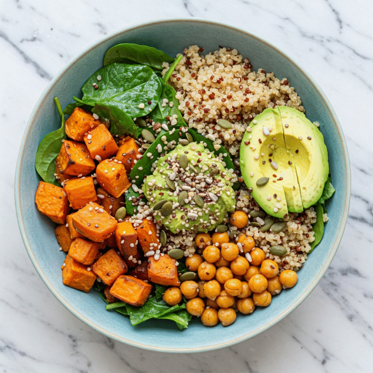 A colorful bowl filled with quinoa, roasted sweet potatoes, chickpeas, avocado, spinach, and seeds on a marble background.