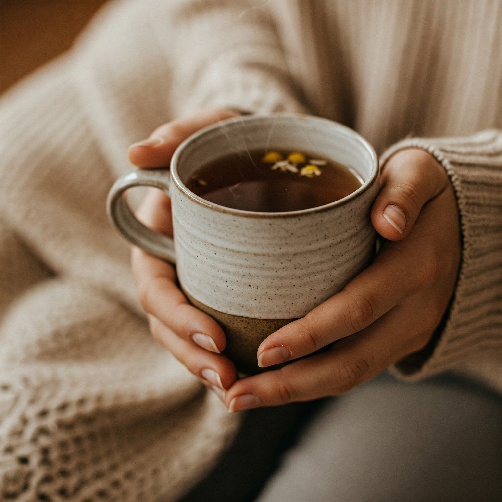 A person wearing a cozy sweater holds a steaming mug filled with a light-colored beverage, possibly tea, with small yellow flowers floating on top.